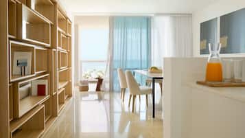 A modern hotel room with a wooden bookshelf, a dining area with a table and chairs, and a view of the sea through a large window.