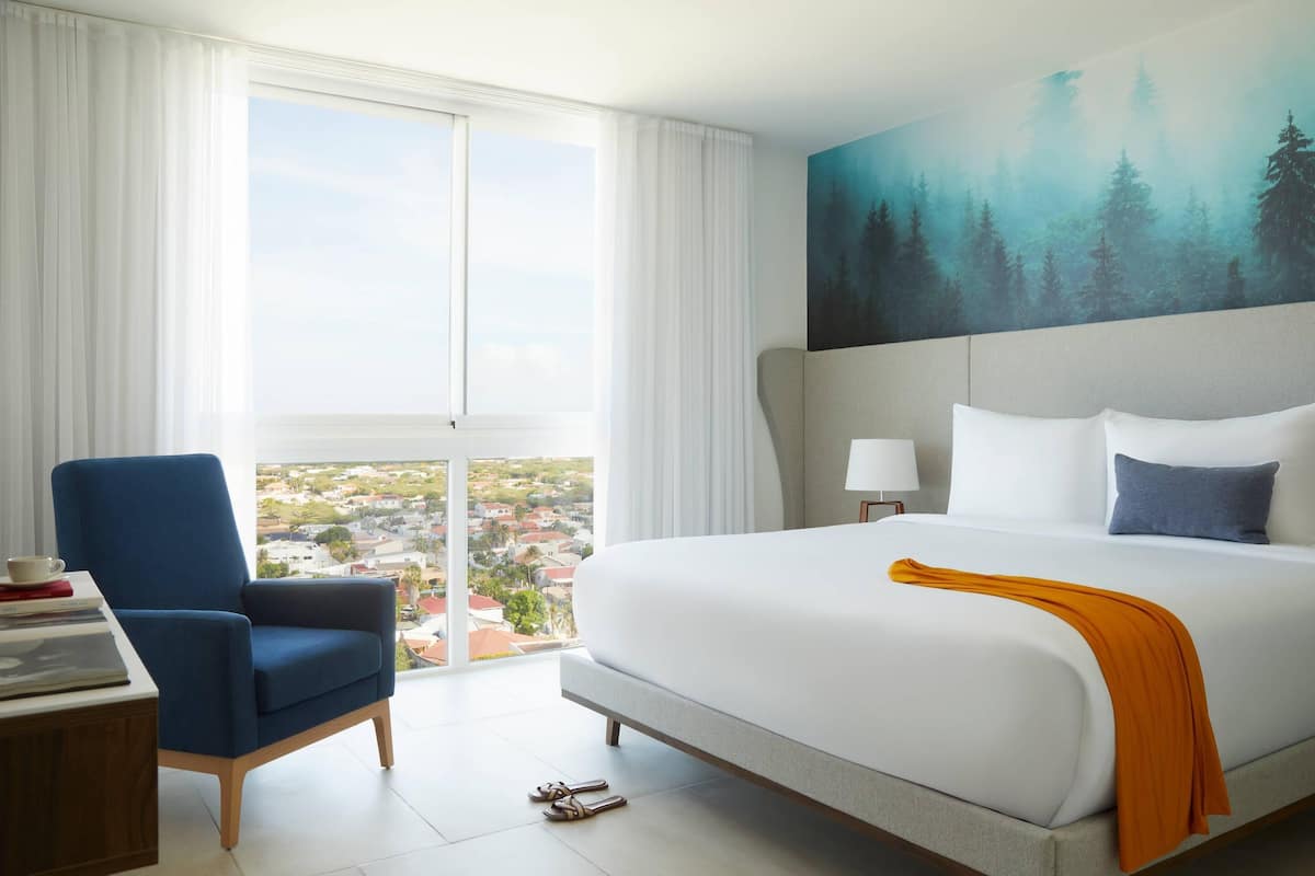 A hotel room with a large bed, a blue armchair, a view of the city through a large window, and a painting of a forest on the wall.