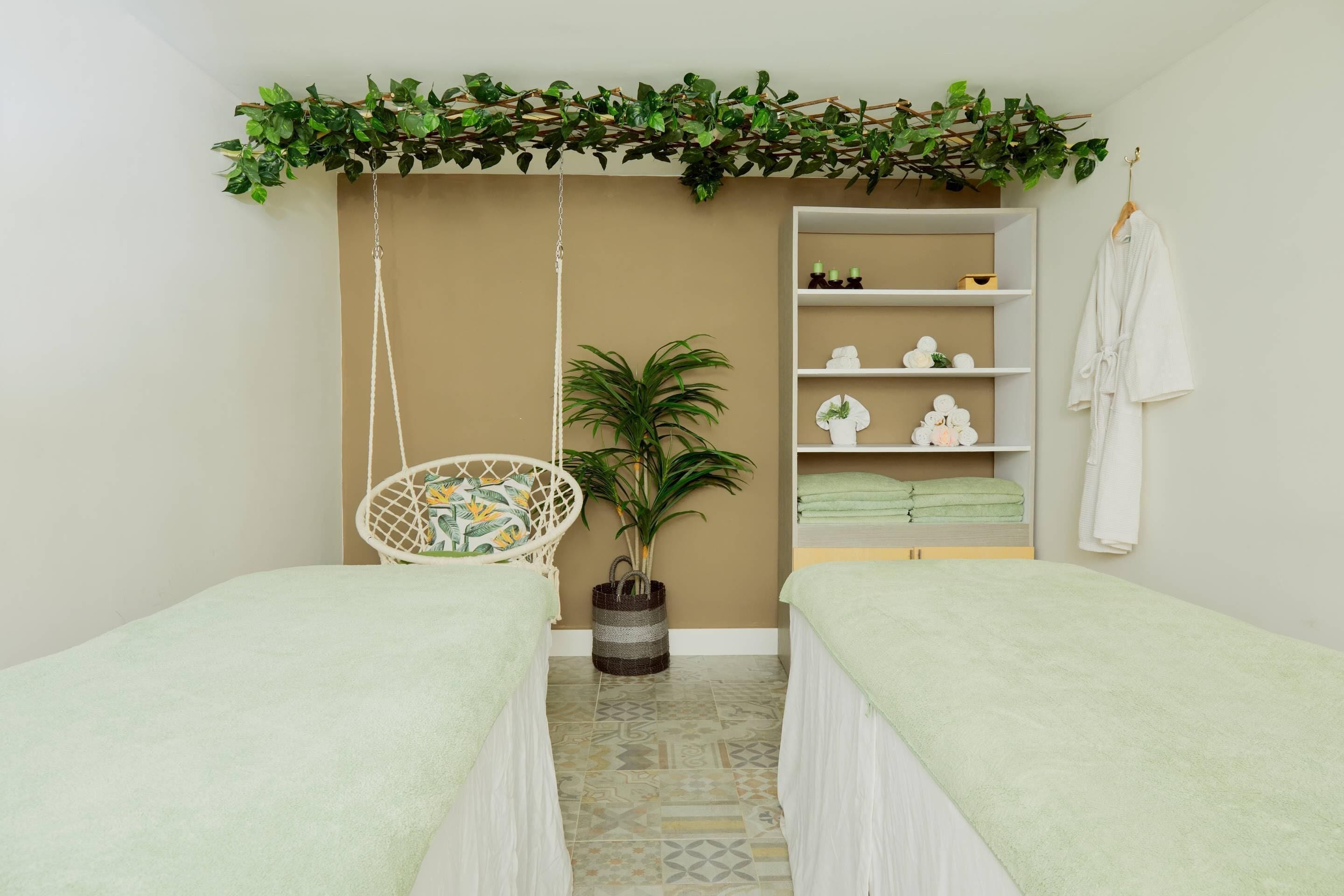 A spa room with two beds, a hanging chair, a potted plant, shelves with toiletries, and a robe hanging on the wall.