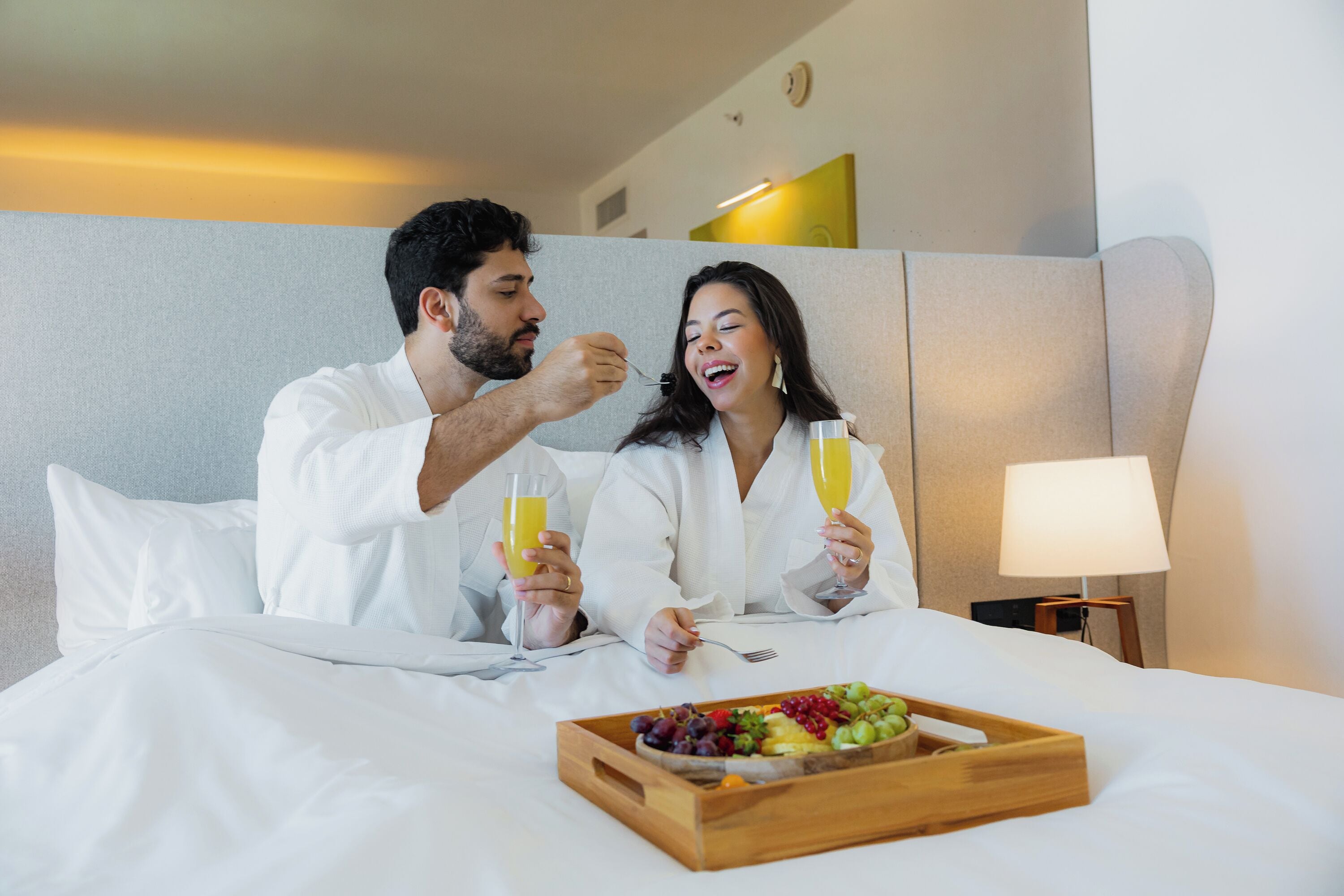 Habitación de hotel con una cama, una bandeja de fruta y dos personas desayunando.