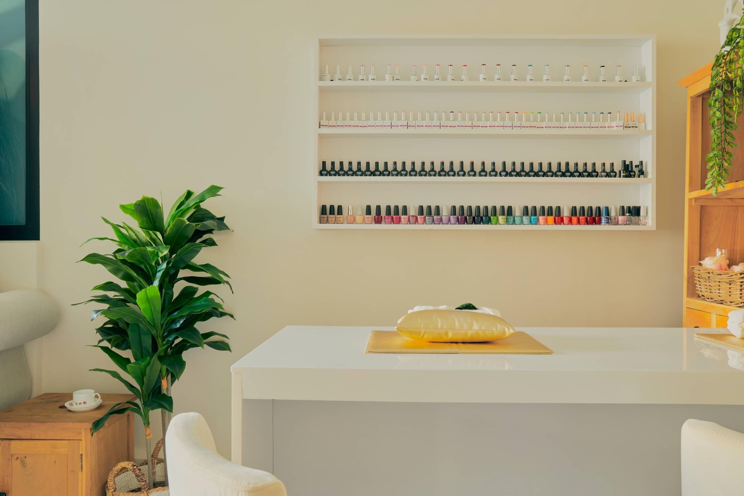A reception area with a white reception desk, a plant, and a wall-mounted shelf with nail polish bottles.
