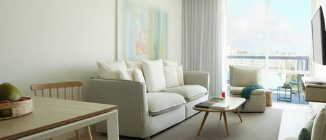 A modern living room with a sofa, a dining table, and a coffee table. There is a window with a view of the city.