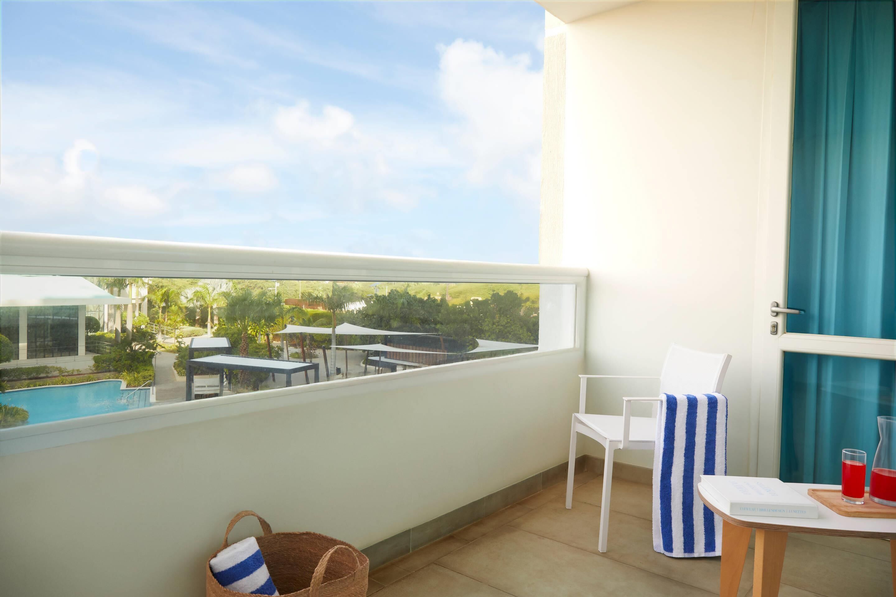 Un balcón con vistas a una piscina y follaje, una silla blanca, una toalla a rayas y una mesa con una bebida y un libro.