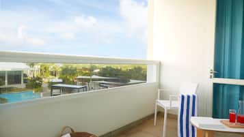 Un balcón con vistas a una piscina y follaje, una silla blanca, una toalla a rayas y una mesa con una bebida y un libro.