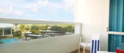 Un balcón con vistas a una piscina y follaje, una silla blanca, una toalla a rayas y una mesa con una bebida y un libro.