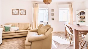 A living room with a beige sofa, a wooden dining table with chairs, and a hanging light fixture. - Caíque – Charming 3-Bed Apartment in Olhão Downtown, Close to Ria Formosa (Olhão)