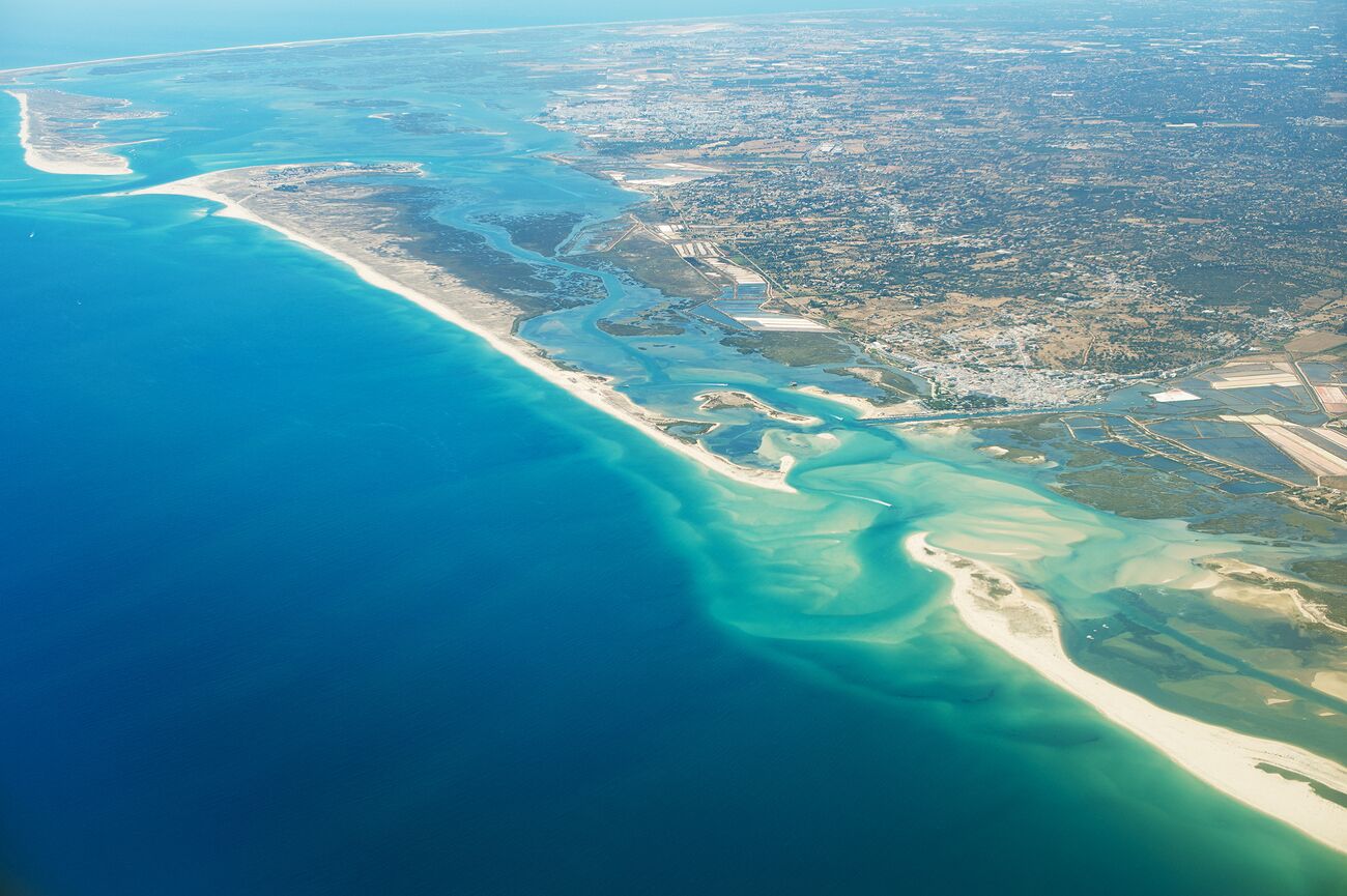 An aerial view of a coastal area with a beach, clear blue waters, and a mix of urban and natural landscapes.
