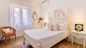 A white bedroom with a four-poster bed, a wicker chair, a bedside table with a lamp, and a wall-mounted air conditioner. - Caíque – Charming 3-Bed Apartment in Olhão Downtown, Close to Ria Formosa (Olhão)
