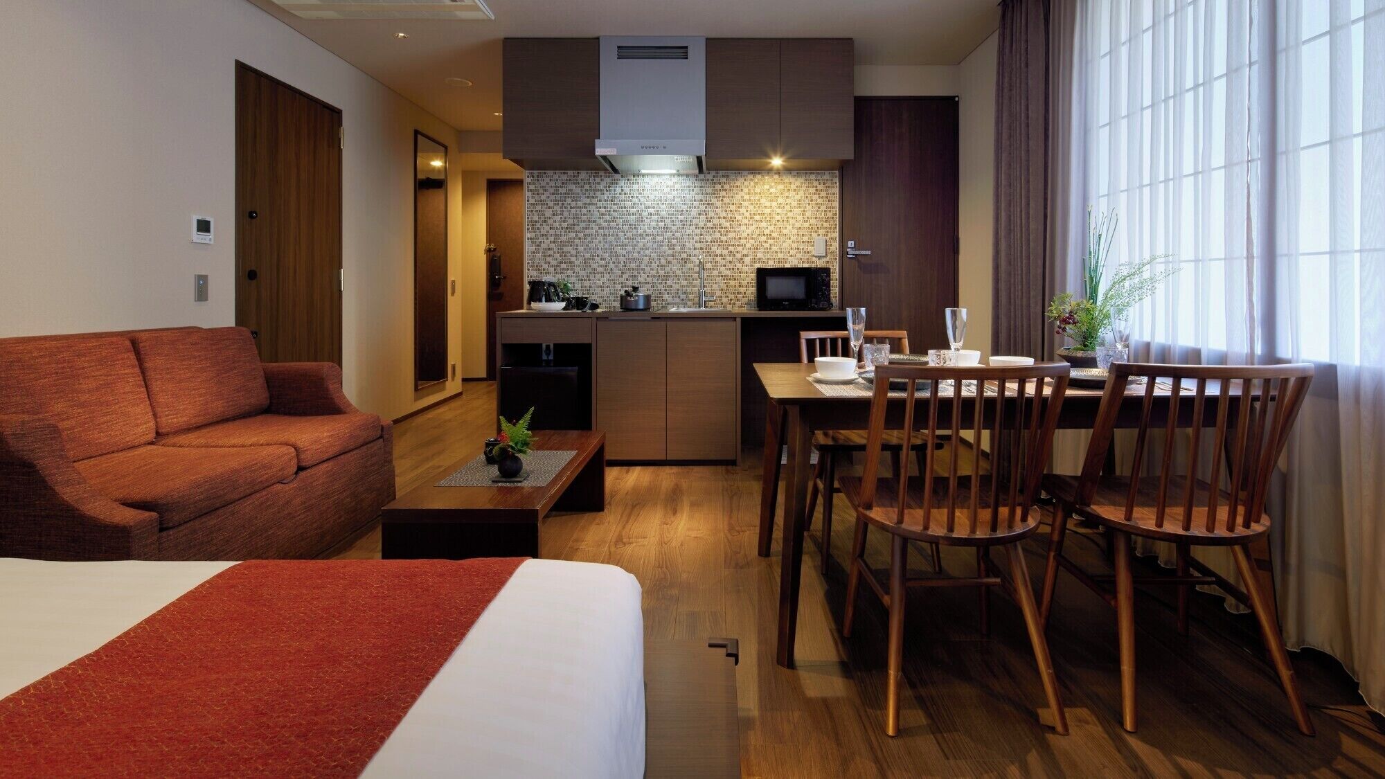 Une chambre d’hôtel compacte comprenant un canapé, une table à manger avec des chaises, un coin cuisine et une grande fenêtre avec des rideaux.