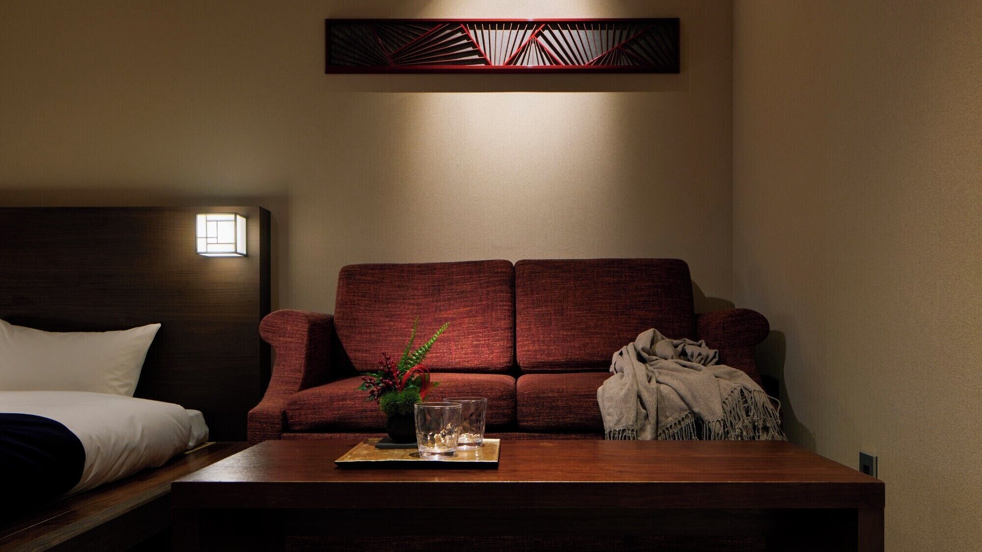 Une chambre d’hôtel avec une tête de lit en bois foncé, un canapé rouge, une table basse en bois sur laquelle se trouvent un verre et une plante, et un luminaire fixé au mur.