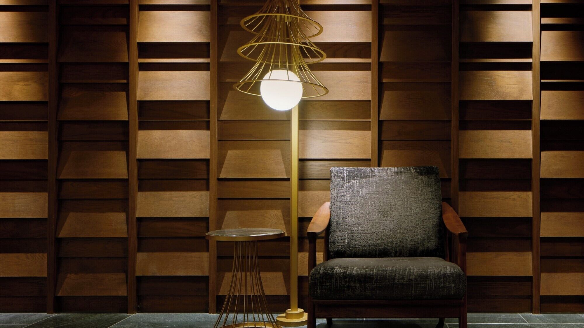 A modern room with a wooden slatted wall, a round side table, a dark upholstered chair, and a unique pendant light.