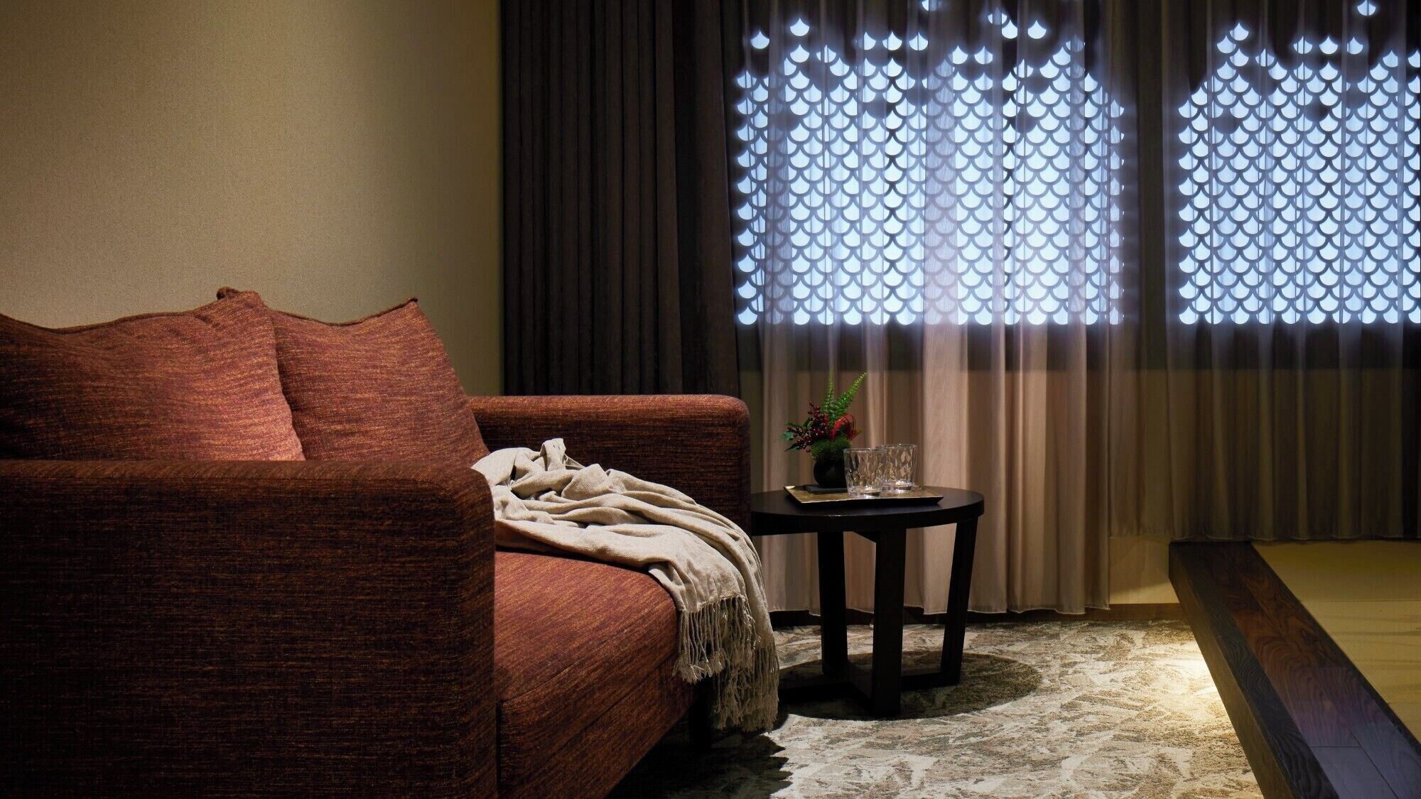 A cozy living room with a brown sofa, a small round table with a plant and glasses, and a window with decorative curtains.
