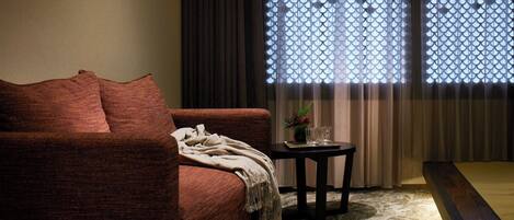 A cozy living room with a brown sofa, a small round table with a plant and glasses, and a window with decorative curtains.