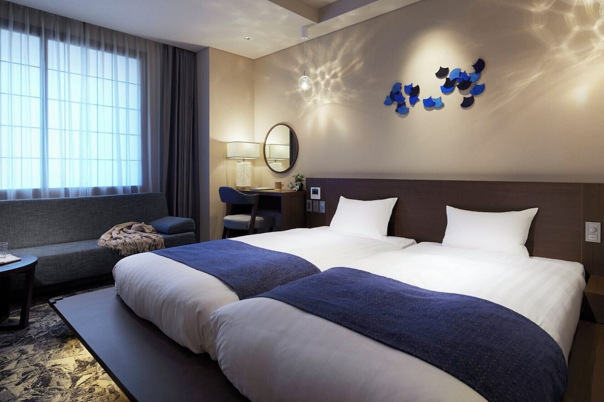Hotelkamer met een groot bed, een bureau, een stoel, een bijzettafeltje met een plant en een raam met gordijnen.