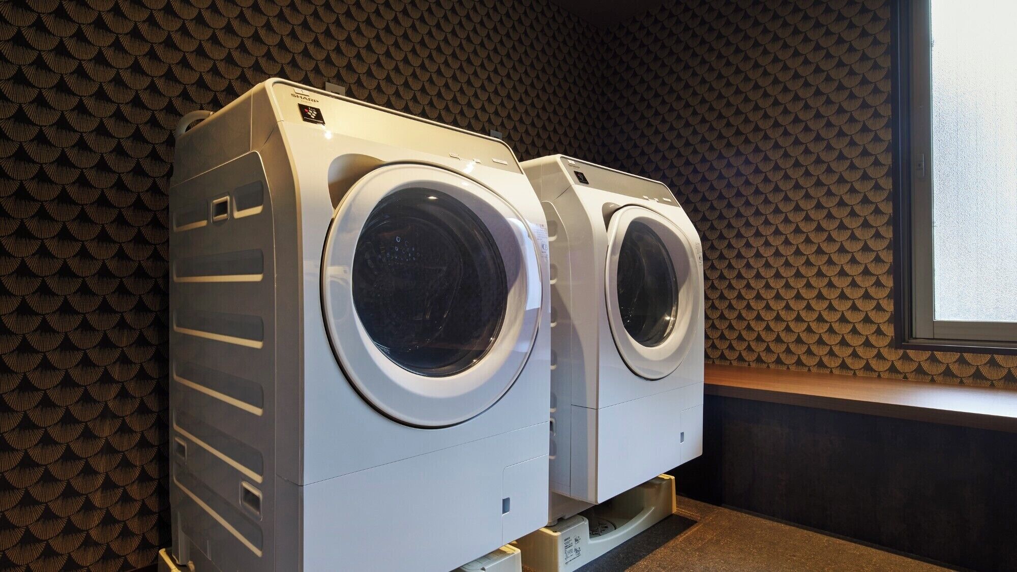 Deux machines à laver à chargement frontal se trouvent dans une buanderie, avec un mur à motifs et une fenêtre.