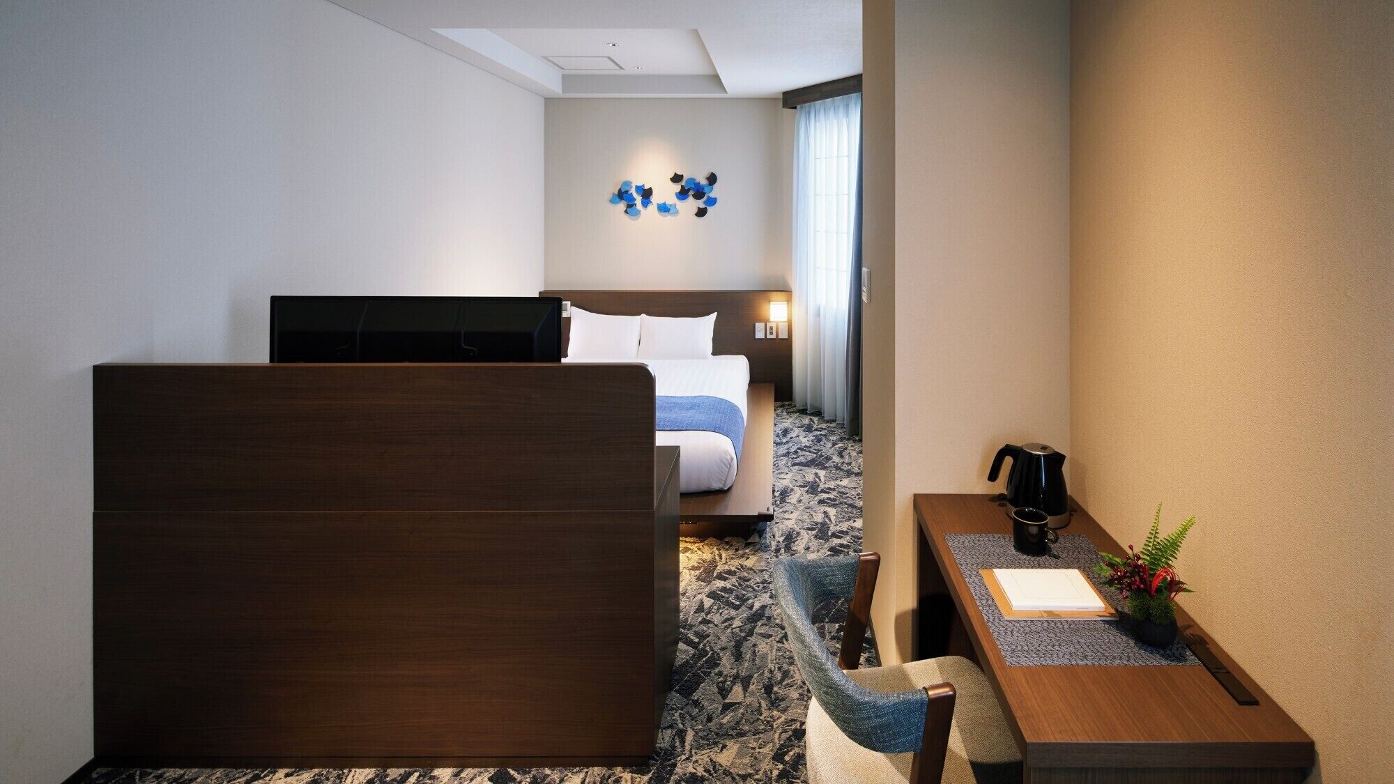 A hotel room with a bed, a desk with a coffee maker, a chair, and a small plant.