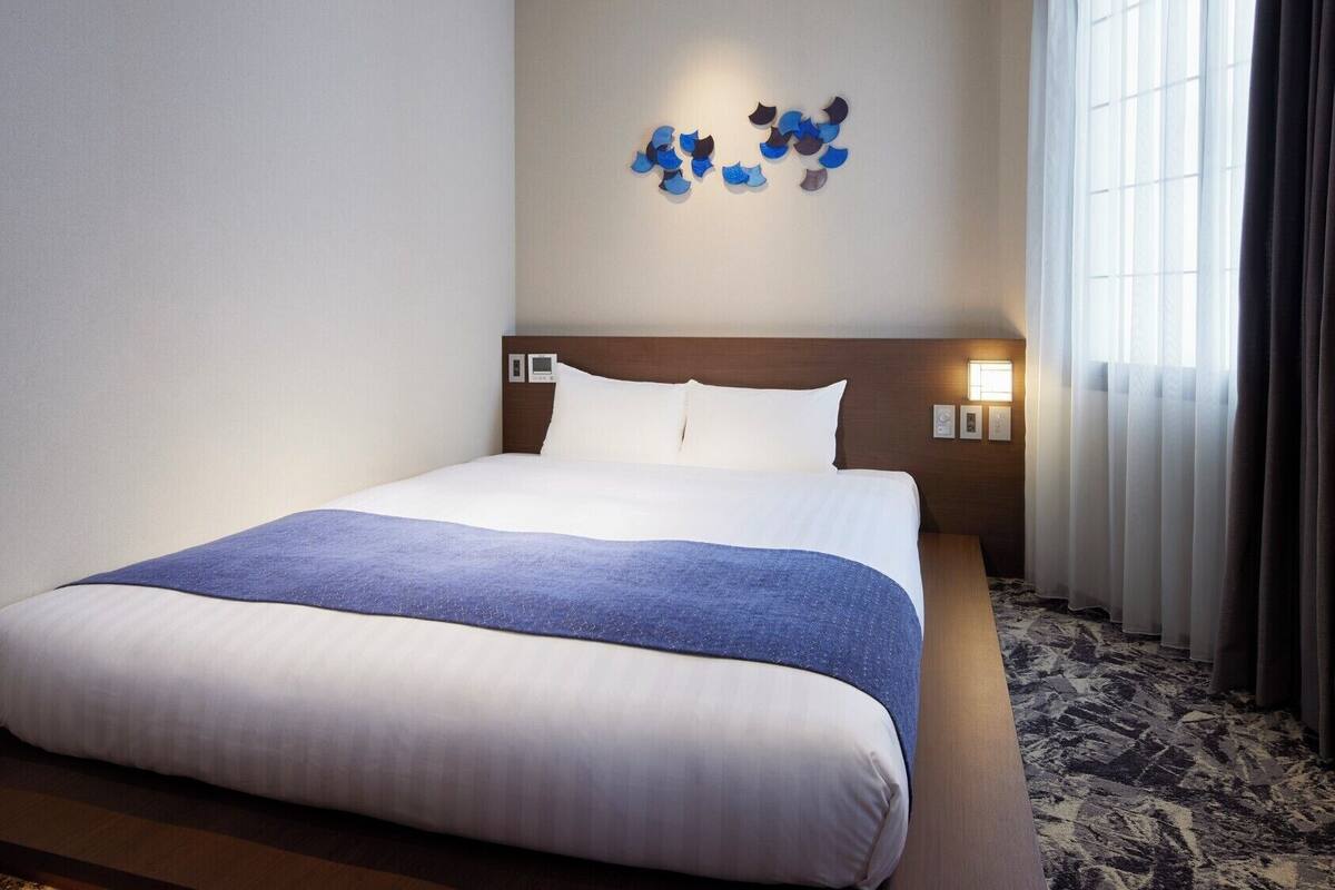 Een hotelkamer met een groot bed, een hoofdeinde met bedieningspaneel, een wandlamp en een raam met gordijnen.