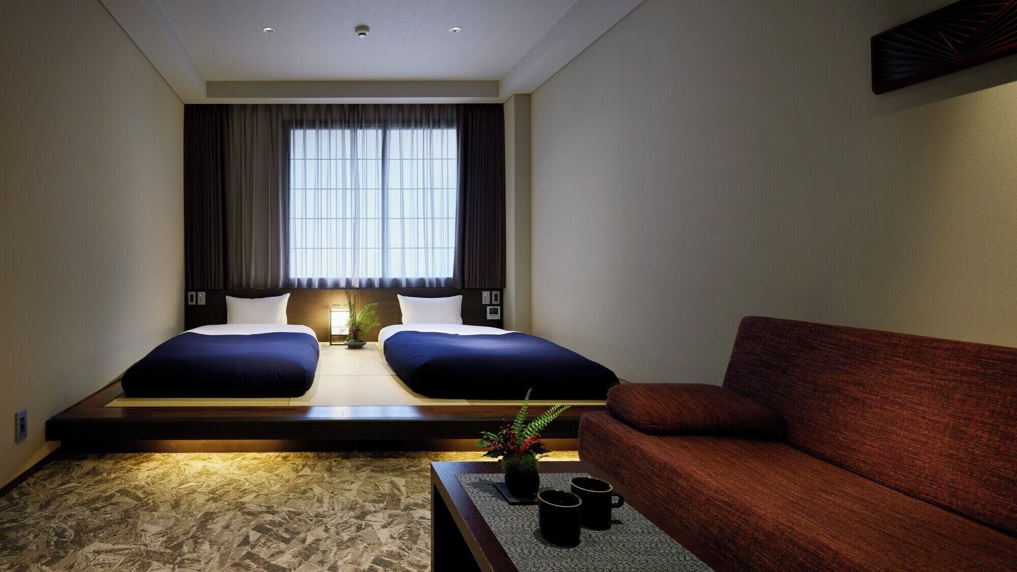 A hotel room with a bed, two pillows, a bedside table with a plant and cups, a sofa, and a window with curtains.