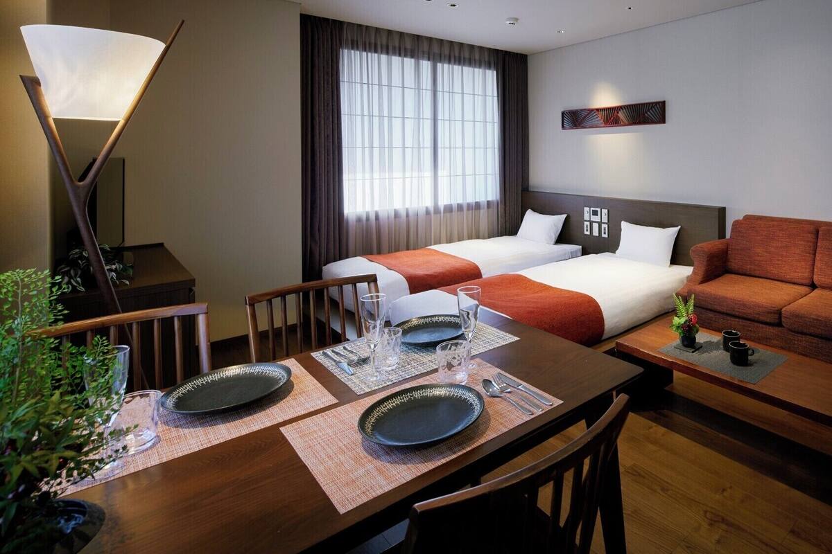 Hotelkamer met twee bedden, een bank, een eettafel gedekt met borden en glazen, een lamp en een groot raam met gordijnen.