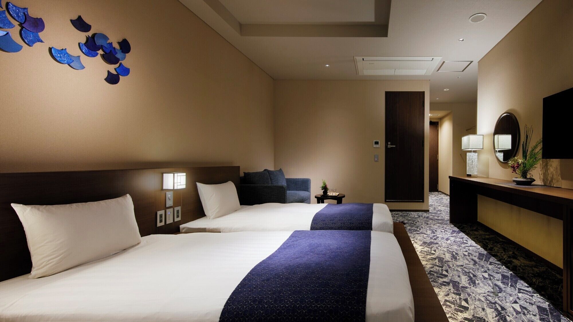 A hotel room with two beds, a desk with a lamp, a mirror, and a decorative wall piece.