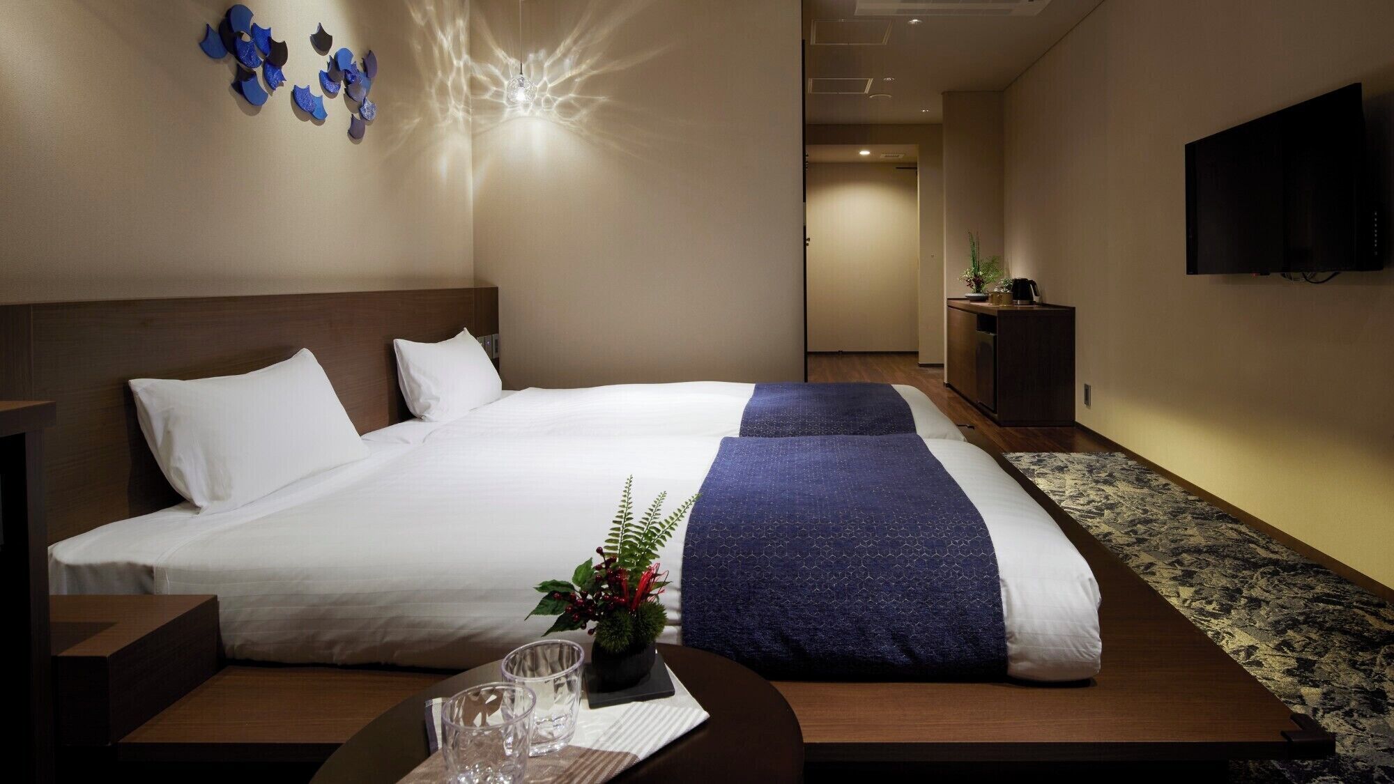Quarto de hotel com uma cama grande, uma escrivaninha com cadeira, uma mesinha com copos e uma planta, e uma televisão fixada na parede.