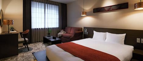 Habitación de hotel con una cama grande, un escritorio, una silla, una mesita, una planta en maceta y una ventana con cortinas.