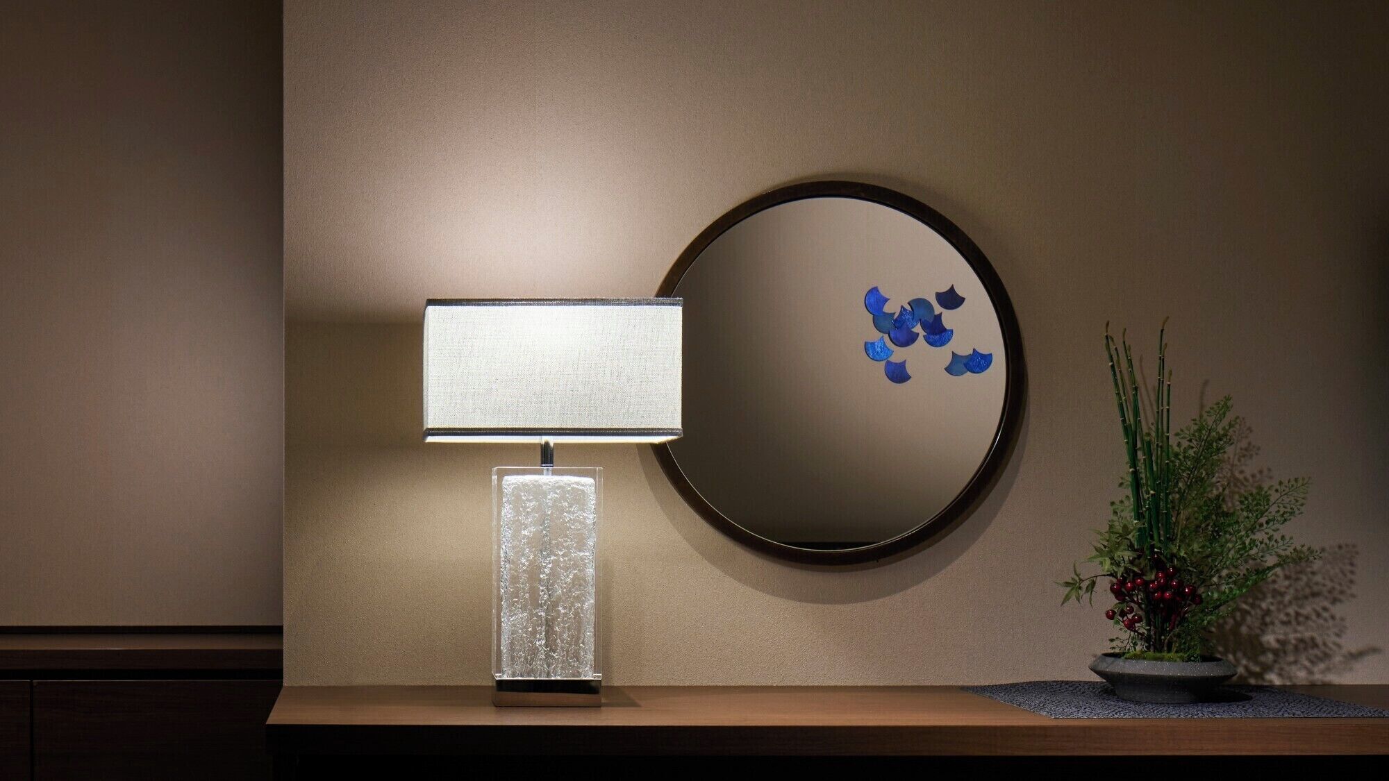 A wall-mounted round mirror with a dark frame, a rectangular table lamp with a textured shade, and a small potted plant with green foliage and red berries.