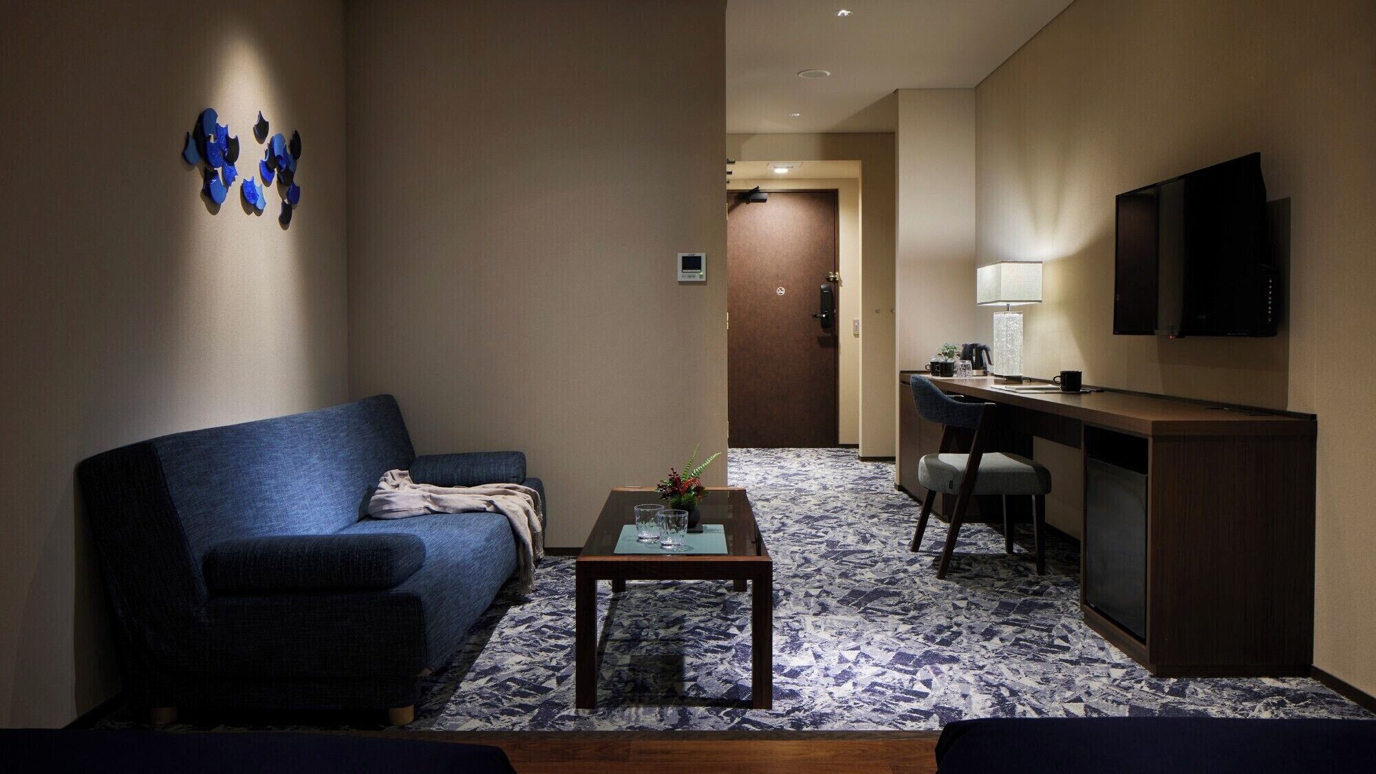 Ein Hotelzimmer mit einem blauen Sofa, einem kleinen Couchtisch, einem Schreibtisch mit Stuhl, einem an der Wand befestigten Fernseher und einem gemusterten Teppich.