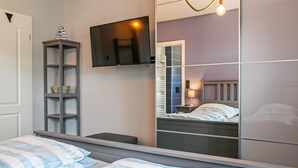 Un dormitorio con cama, mesita de noche, espejo, televisor montado en la pared y estantería.