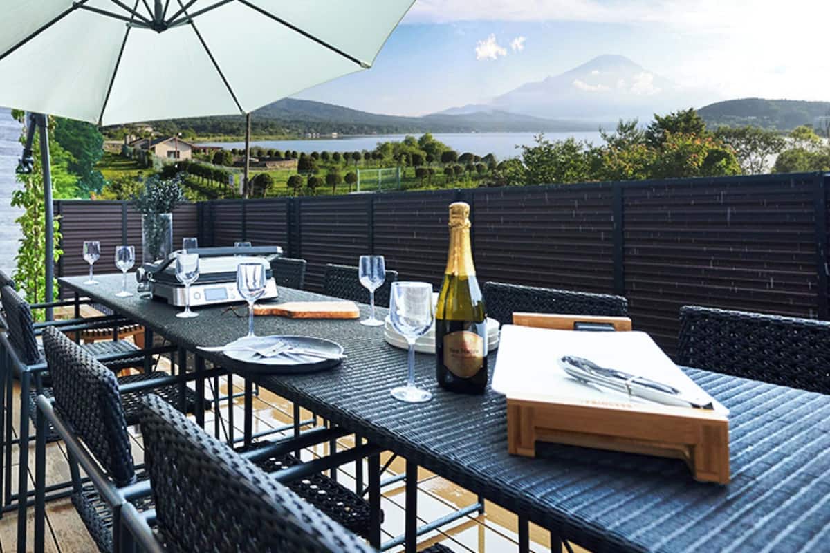Un área de comedor al aire libre con vistas a un lago y montañas, equipada con una parrilla, mesa, sillas y una sombrilla.