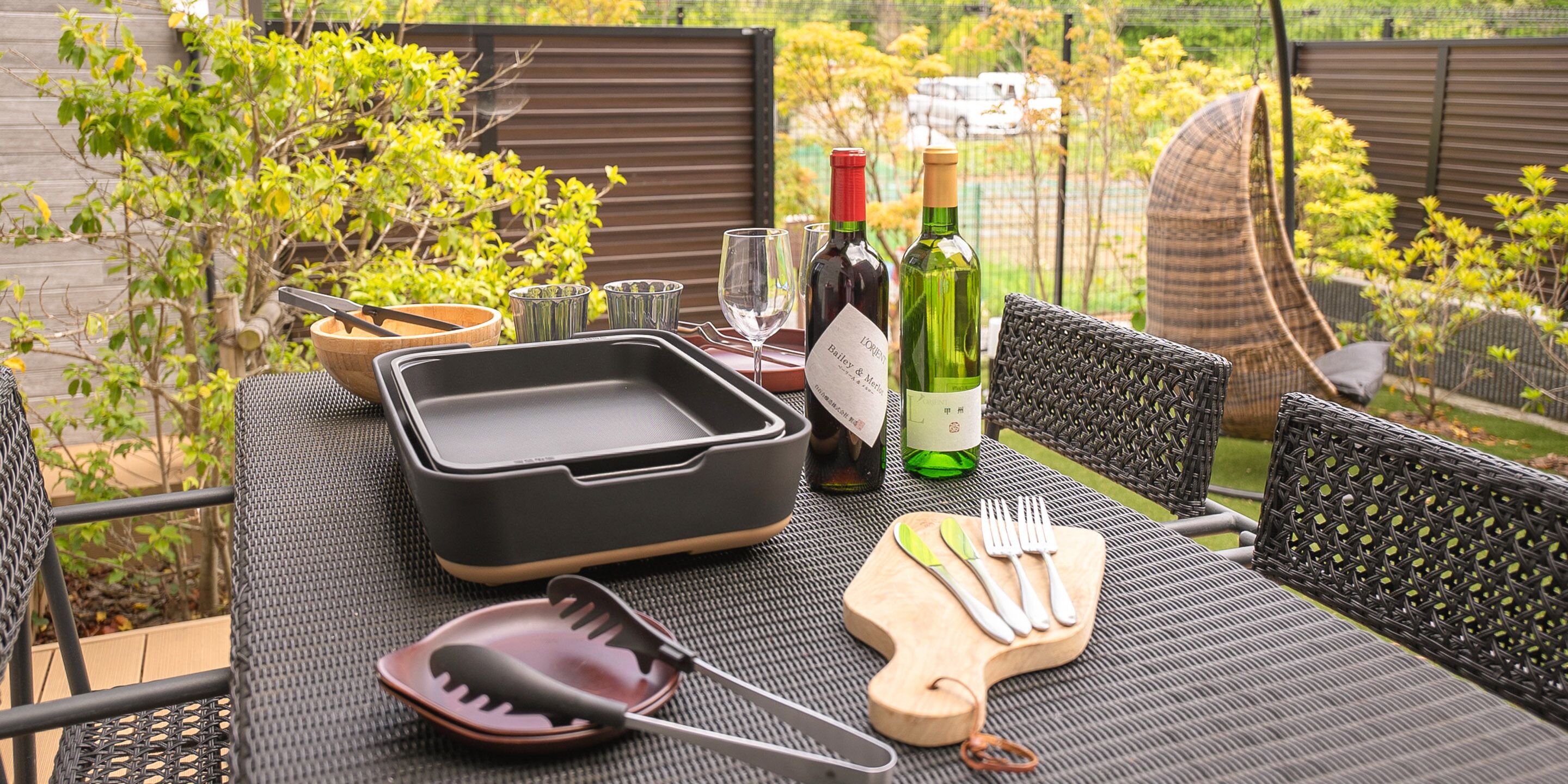 Une table à manger installée à l’extérieur, agrémentée d’un plateau noir, de bouteilles de vin, de couverts et d’une chaise en osier.