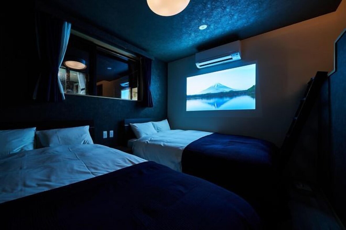 A dimly lit room with two beds, a large window, and a projection screen displaying a scenic landscape.