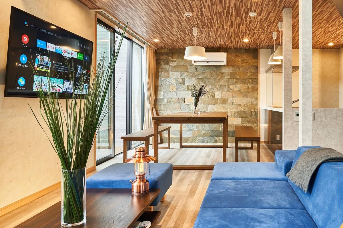 Una sala de estar moderna con un sofá azul, una mesa de comedor de madera y un televisor montado que muestra servicios de transmisión.