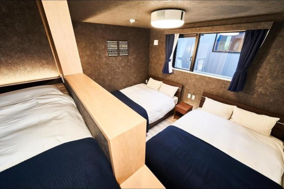 A compact hotel room with two beds, a small window with curtains, and a wall-mounted light fixture.