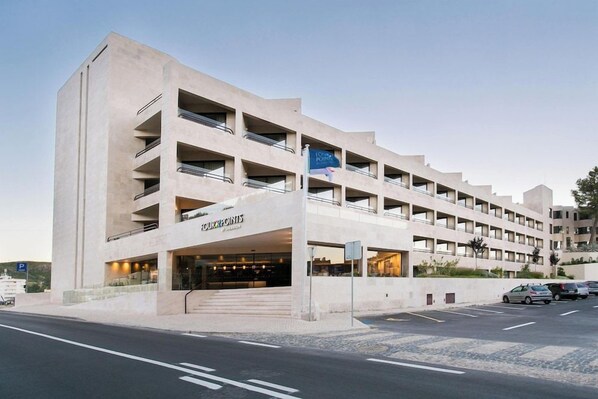 A multi-story hotel with a modern design, featuring a prominent entrance and a parking area in front.