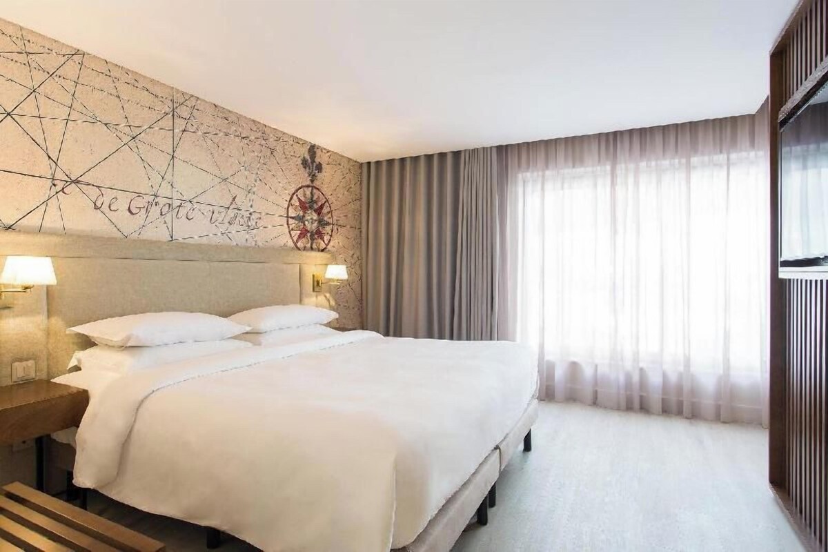 Hotellihuone, jossa on suuri sänky, kaksi yöpöytävalaisinta, koristeellinen seinä, jossa on kompassi, ja suuri ikkuna, jossa on verhot.