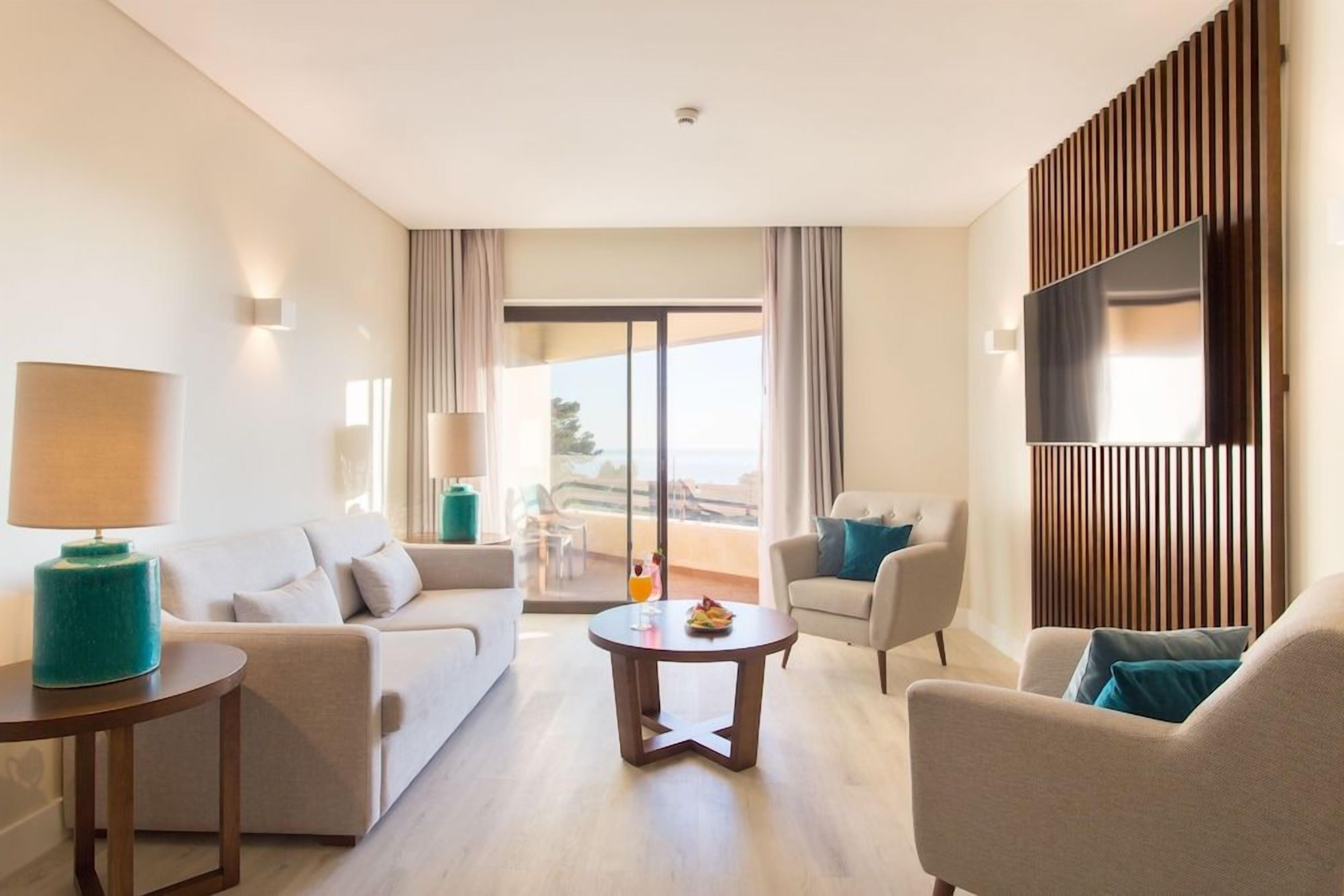 A modern hotel room with a sofa, armchairs, a coffee table, a TV, and a balcony with a view.