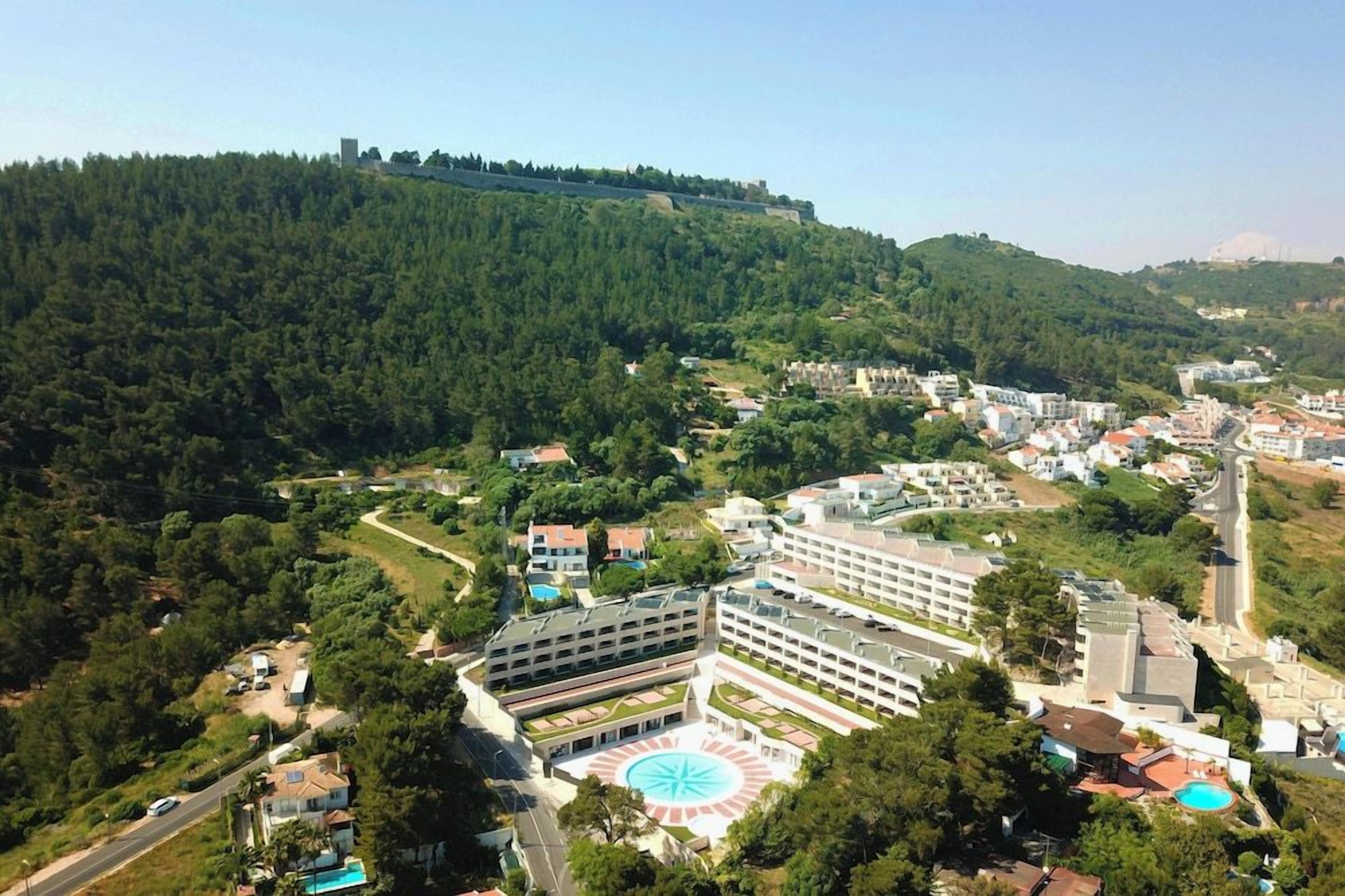 A hotel with multiple buildings, a central courtyard with a circular design, and a surrounding forested area.