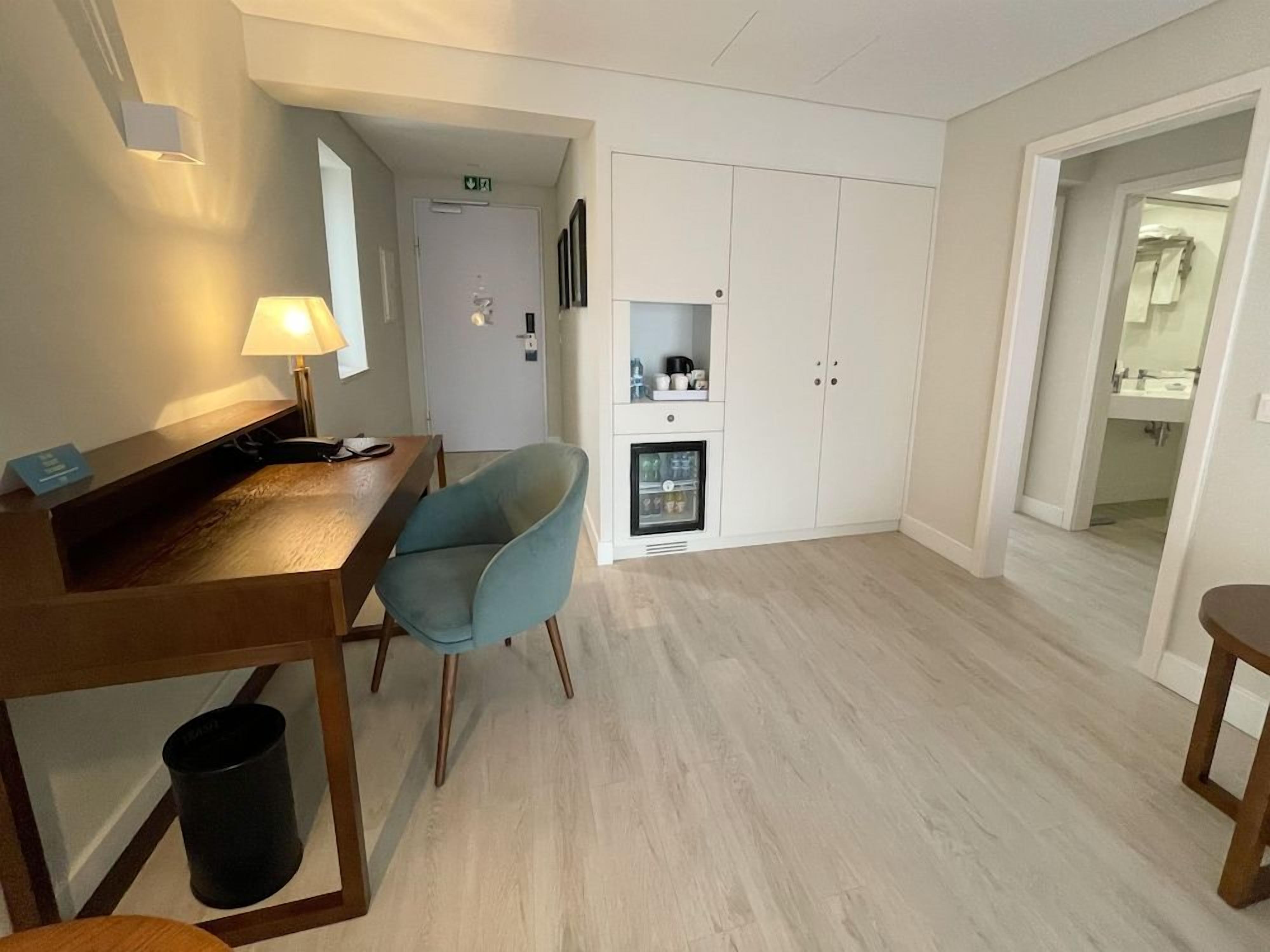 A hotel room with a wooden desk, a blue chair, a wardrobe, and a small kitchenette.