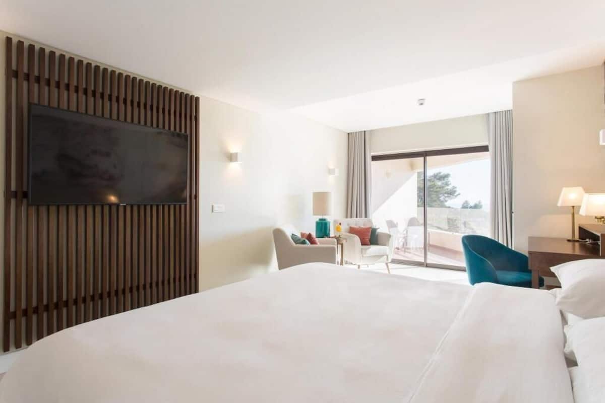 A hotel room with a large bed, a flat-screen TV mounted on a wooden panel, a seating area with chairs, and a view through a large window.