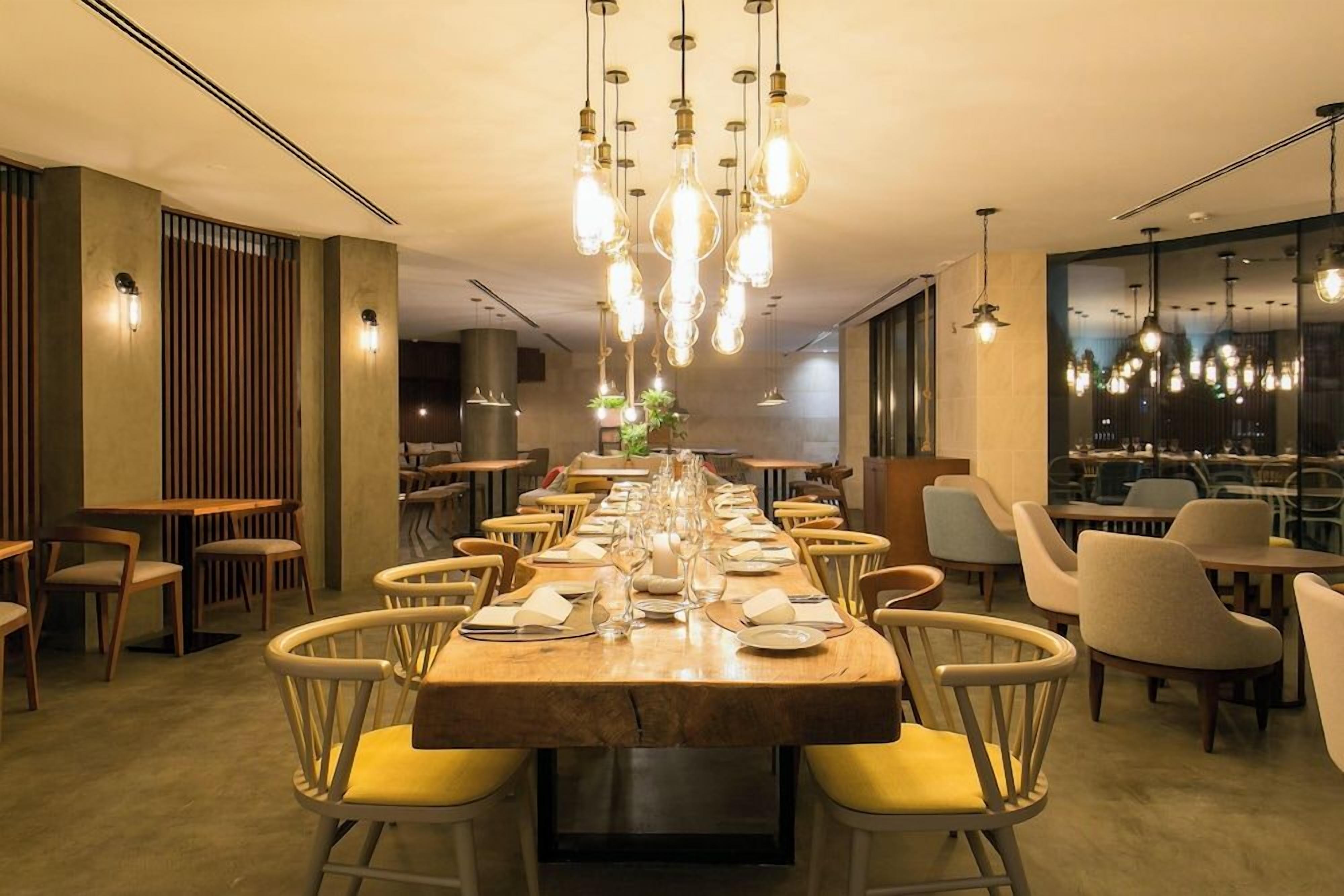 A restaurant with a long communal table set for dining, surrounded by chairs, and hanging pendant lights.