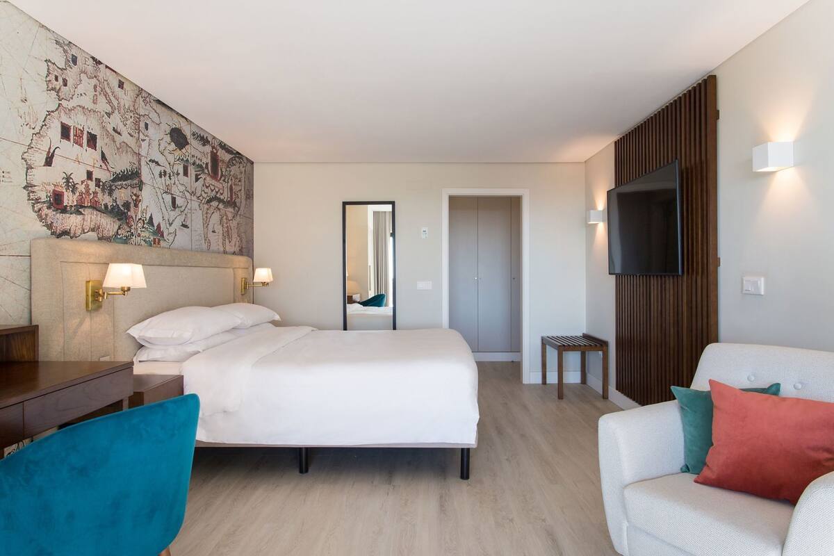 A hotel room with a large bed, a desk, a chair, a TV, and a mirror.
