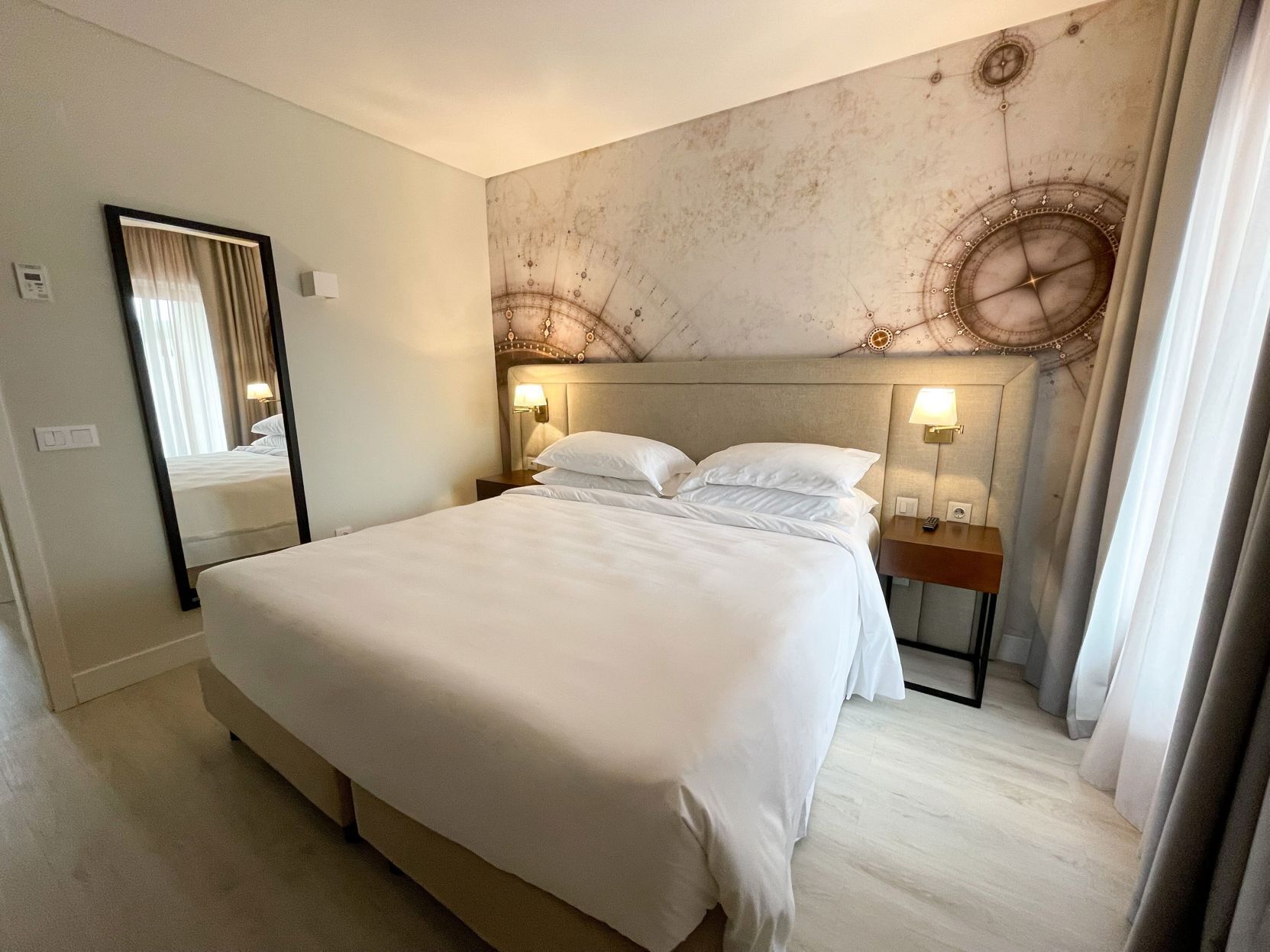 Et hotelværelse med en stor seng, et spejl, to sengeborde med lamper og en væg med dekorativt design.