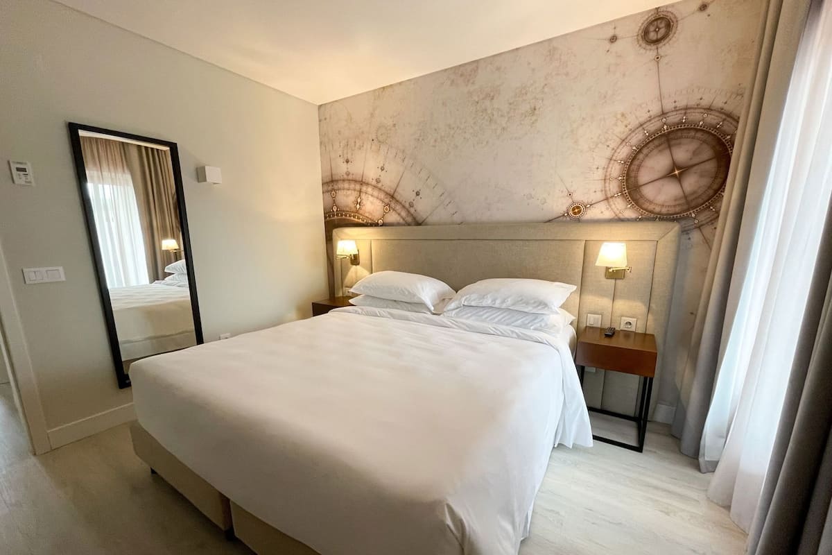 A hotel room with a large bed, a mirror, two bedside tables with lamps, and a wall with a decorative design.