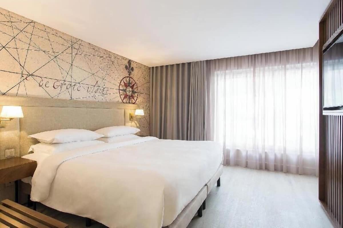 A hotel room with a large bed, two bedside lamps, a decorative wall with a compass, and a large window with curtains.