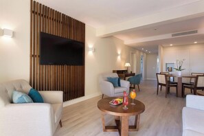 A modern living room with a flat-screen TV, a round wooden coffee table with fruit and drinks, a sofa, and a dining area with a table and chairs.