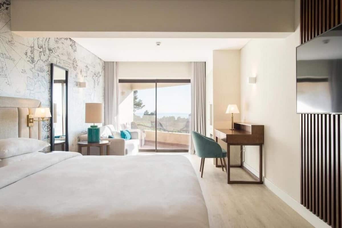 A hotel room with a large bed, a desk with a chair, a TV, and a balcony with a view.