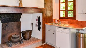 A kitchen with a sink, a dish rack, a white dishwasher, a window with a view of greenery, and a rustic brick fireplace.