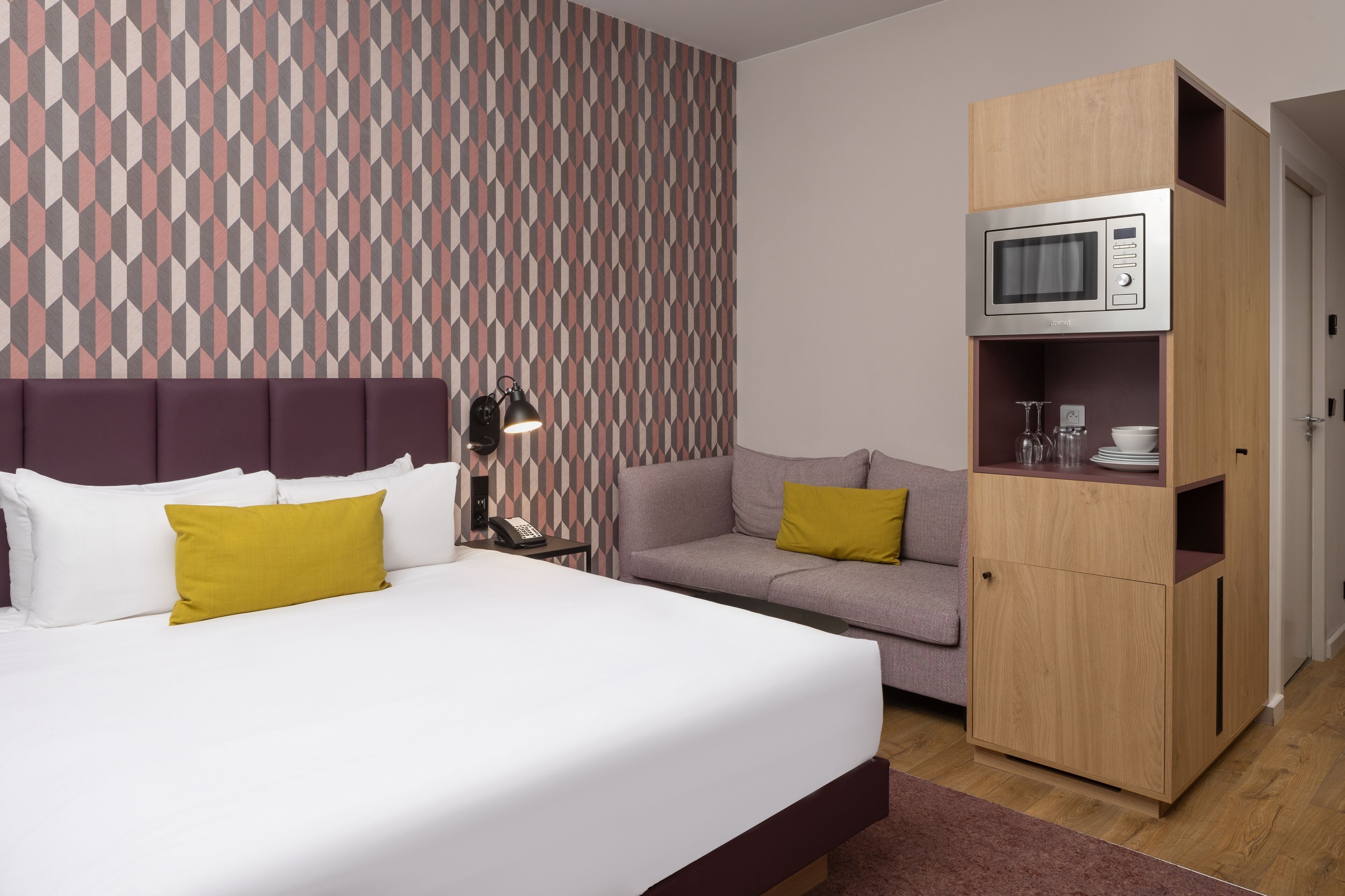 Habitación de hotel con una cama, un sofá, un mueble de madera con microondas y una pared con diseño.