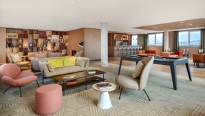 Una sala moderna con un sofá, sillones, una mesa baja y una mesa de billar. Se ve una estantería y una zona de cocina al fondo.